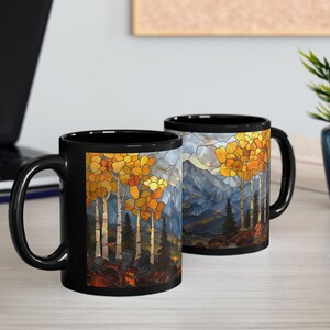 May include: Two black ceramic mugs with a stained glass design of a mountain landscape with trees in fall colors. The mugs are on a white surface.