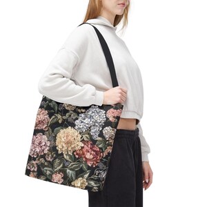 May include: Black tote bag with a floral pattern in pink, cream, and blue. The bag has a black strap and is held by a person. The floral design features peonies, hydrangeas, and other flowers.