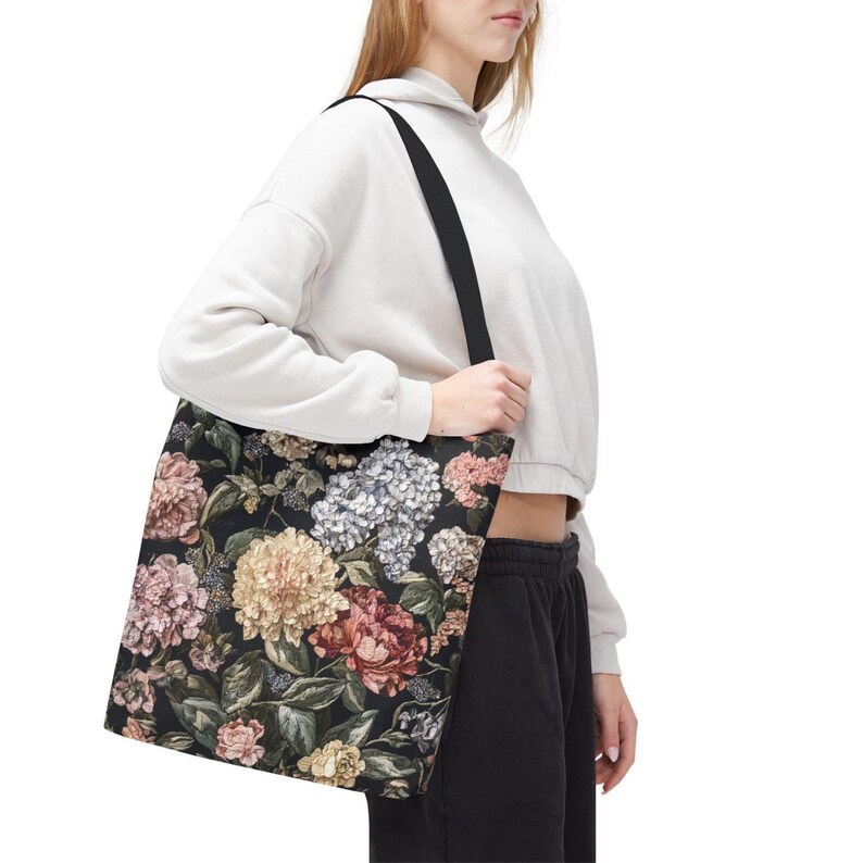 May include: Black tote bag with a floral pattern in pink, cream, and blue. The bag has a black strap and is held by a person. The floral design features peonies, hydrangeas, and other flowers.