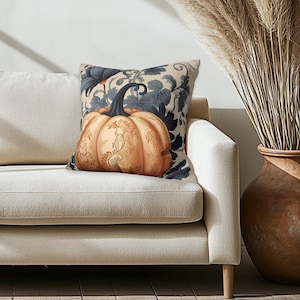 May include: A square throw pillow featuring a central orange pumpkin with intricate gold filigree patterns. The pumpkin has a dark stem and is set against a background of dark blue botanical leaves and vines on a light cream-colored fabric base.
