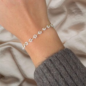 Op de afbeelding: Een delicate gouden kettingarmband met kleine witte bloemvormige kralen. De armband zit om een pols.