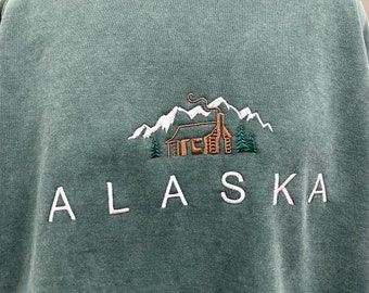 Könnte beinhalten: Ein grüner Pullover mit einem weißen gestickten Design einer Hütte in den Bergen mit dem Wort "ALASKA" darunter.