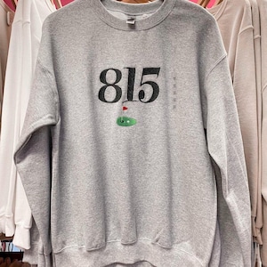 May include: Gray sweatshirt with a black embroidered design of the number 815 and a golf green with a red flag.