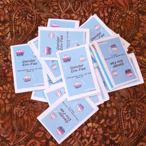 May include: A stack of blue cards with a pink and purple flower design. The cards have the text "Gender Env-Fae (Like gender envy, but fae) haha :D" printed on them.