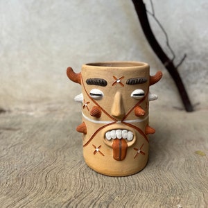 May include: A terracotta planter shaped like a face with horns, white eyes, and a red tongue sticking out. The face has a white cross on the forehead and white X's on the cheeks.