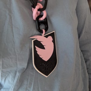 K&ouml;nnte beinhalten: Eine auff&auml;llige Halskette mit rosa und schwarzen Kettengliedern. Der Anh&auml;nger ist eine schwarze Schildform mit einer rosa Taube und dem Text "ANGEL CITY FC".