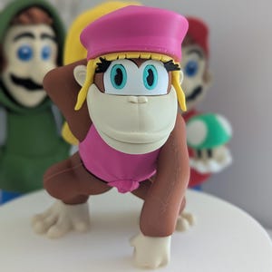 Pode incluir: Uma figura de macaco fêmea impressa em 3D de cor marrom e rosa, usando um chapéu rosa e um maiô rosa. O macaco tem olhos azuis e cabelo loiro.