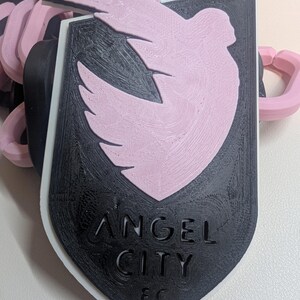 K&ouml;nnte beinhalten: Ein 3D-gedruckter Schl&uuml;sselanh&auml;nger mit dem Logo von Angel City FC. Das Design umfasst ein schwarzes Schild mit dem Teamnamen und einem rosa Taubenemblem. Der Schl&uuml;sselanh&auml;nger hat schwarze und rosa Kettenglieder.