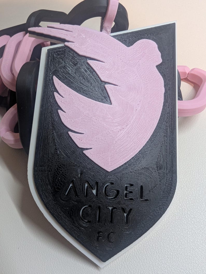 K&ouml;nnte beinhalten: Ein 3D-gedruckter Schl&uuml;sselanh&auml;nger mit dem Logo von Angel City FC. Das Design umfasst ein schwarzes Schild mit dem Teamnamen und einem rosa Taubenemblem. Der Schl&uuml;sselanh&auml;nger hat schwarze und rosa Kettenglieder.