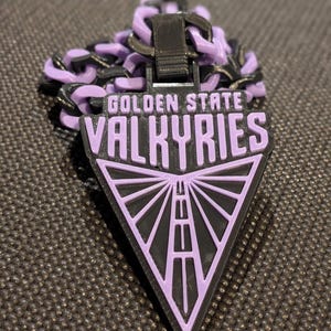 May include: A black and light purple plastic chain necklace featuring a triangular pendant. The pendant displays the text GOLDEN STATE VALKYRIES in raised lettering above a stylized road graphic that converges toward the bottom point.