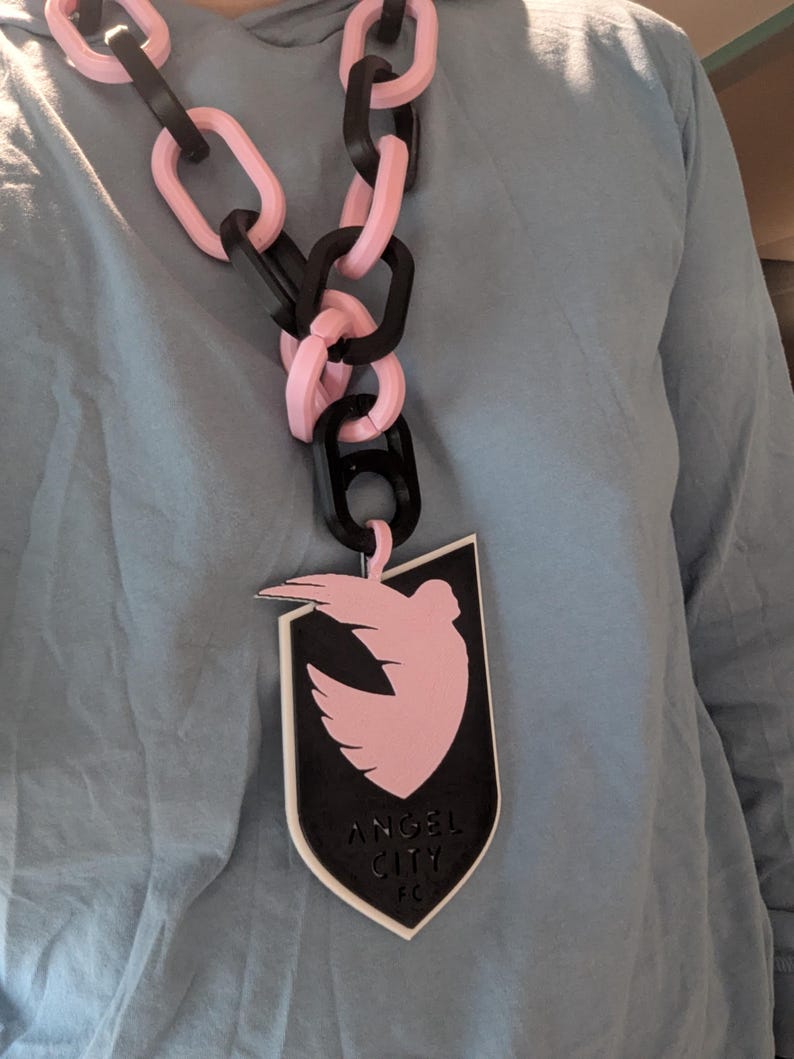 K&ouml;nnte beinhalten: Eine auff&auml;llige Halskette mit rosa und schwarzen Kettengliedern. Der Anh&auml;nger ist eine schwarze Schildform mit einer rosa Taube und dem Text "ANGEL CITY FC".