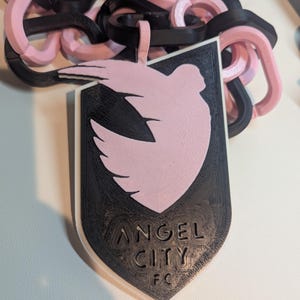K&ouml;nnte beinhalten: Ein schwarz-rosa Anh&auml;nger mit einem Taubendesign und dem Text "ANGEL CITY FC". Der Anh&auml;nger ist mit einer Kette aus ineinandergreifenden Gliedern in Schwarz und Rosa verbunden. Das Design ist 3D-gedruckt.