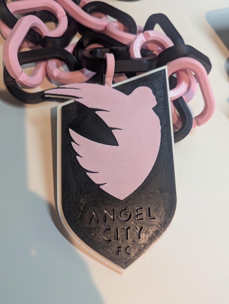 K&ouml;nnte beinhalten: Ein schwarz-rosa Anh&auml;nger mit einem Taubendesign und dem Text "ANGEL CITY FC". Der Anh&auml;nger ist mit einer Kette aus ineinandergreifenden Gliedern in Schwarz und Rosa verbunden. Das Design ist 3D-gedruckt.