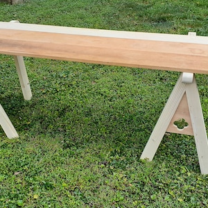 May include: A wooden sawhorse table with a light-colored top and two A-frame supports. The tabletop is made of three different wood tones. The supports are painted a light green color and have a decorative cutout.