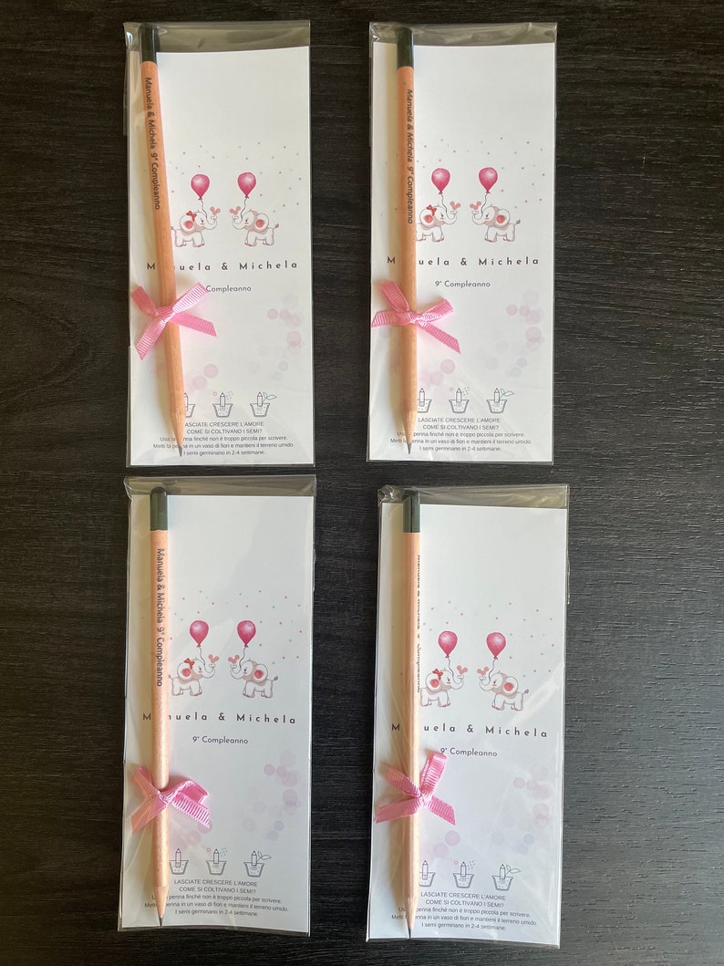 May include: Four white cards with pink and black illustrations of elephants holding balloons. Each card has a pink ribbon and a wooden pencil with a black eraser. The text on the cards reads "Manuela & Michela 9 Compleanno" and "Muela & Michela Compleanno".