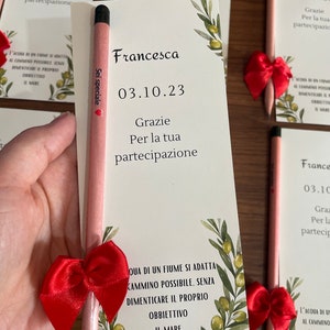 Op de afbeelding: Een roze potlood met een rode strik eromheen. Op het potlood staat "Sei speciale". Het potlood ligt op een witte kaart met groene olijftakken en de tekst "Francesca 03.10.23 Grazie Per la tua partecipazione" erop.
