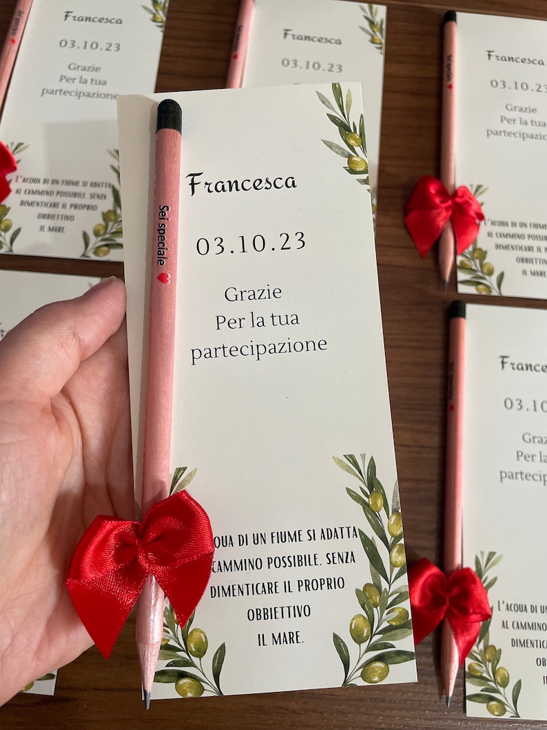 Op de afbeelding: Een roze potlood met een rode strik eromheen. Op het potlood staat "Sei speciale". Het potlood ligt op een witte kaart met groene olijftakken en de tekst "Francesca 03.10.23 Grazie Per la tua partecipazione" erop.