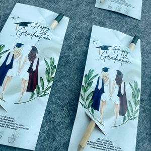 Może przedstawiać: Dwie postacie w togach i czapkach absolwent&oacute;w stoją plecami do siebie. Karta zawiera tekst "Happy Graduation" i "Let Love Grow" z instrukcjami dotyczącymi sadzenia nasion. Karta zawiera drewniany oł&oacute;wek, kt&oacute;ry jest opakowaniem nasion.