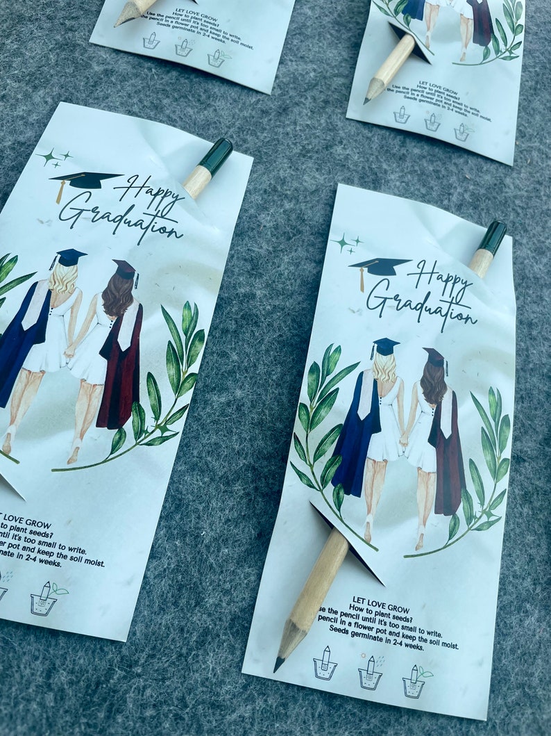 Może przedstawiać: Dwie postacie w togach i czapkach absolwent&oacute;w stoją plecami do siebie. Karta zawiera tekst "Happy Graduation" i "Let Love Grow" z instrukcjami dotyczącymi sadzenia nasion. Karta zawiera drewniany oł&oacute;wek, kt&oacute;ry jest opakowaniem nasion.