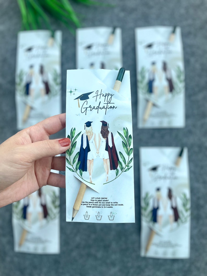 Może przedstawiać: Biała kartka z tekstem "Happy Graduation" i rysunkiem dw&oacute;ch kobiet w togach absolwent&oacute;w. Kartka jest przymocowana do drewnianego oł&oacute;wka z zielonym końc&oacute;wką. Na kartce znajduje się tekst "Let Love Grow" wraz z instrukcjami, jak posadzić oł&oacute;wek, aby wyrosły kwiaty.