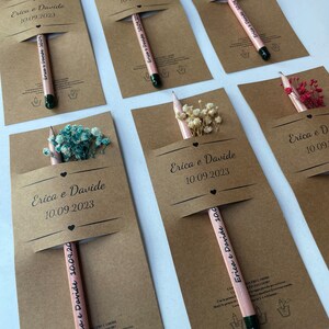 May include: A set of six brown cardstock favor boxes with a pencil and dried flowers inside. Each box has a white label with the text "Erica e Davide" and the date "10.09.2023".