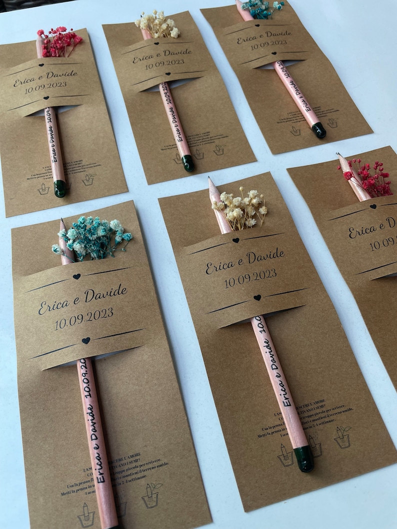 May include: A set of six brown cardstock favor boxes with a pencil and dried flowers inside. Each box has a white label with the text "Erica e Davide" and the date "10.09.2023".
