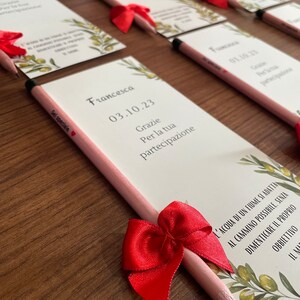 Op de afbeelding: Een roze en witte kaart met de tekst "Francesca 03.10.23 Grazie Per la tua partecipazione" en een rode strik om een roze potlood.