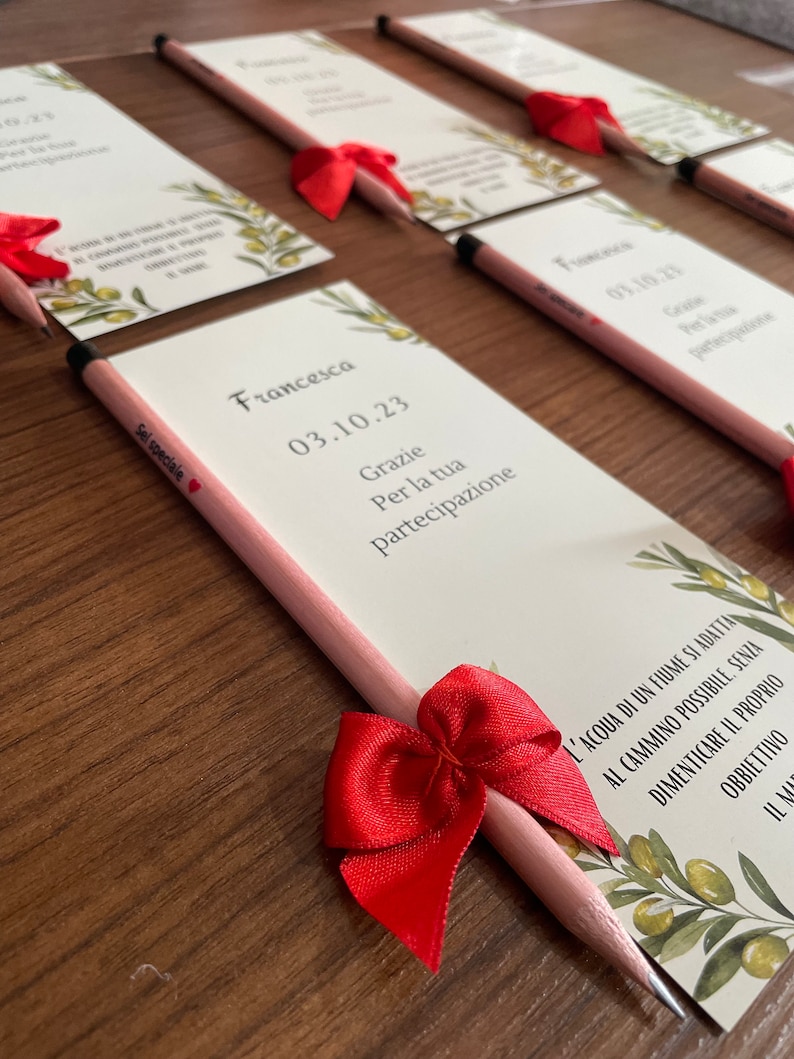 Op de afbeelding: Een roze en witte kaart met de tekst "Francesca 03.10.23 Grazie Per la tua partecipazione" en een rode strik om een roze potlood.