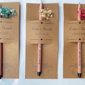 May include: Three kraft paper favor boxes with pink pencils and dried flowers. Each box has a label with the text "Erica e Davide" and the date "10.09.2023".