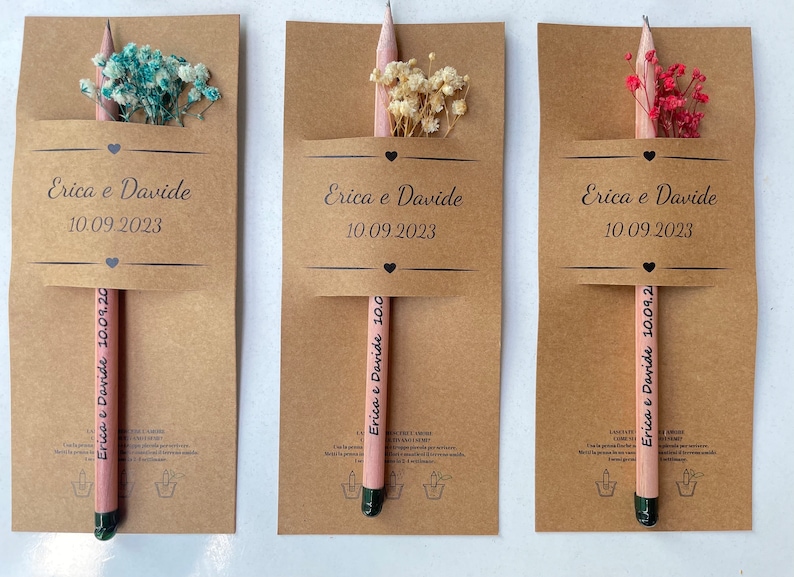 May include: Three kraft paper favor boxes with pink pencils and dried flowers. Each box has a label with the text "Erica e Davide" and the date "10.09.2023".