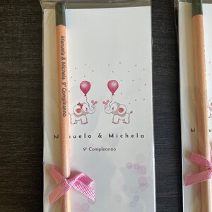 May include: A pink and white card with two elephants holding balloons. The card says "Manuela & Michela 9th Birthday". A wooden pencil with a pink ribbon is attached to the card.