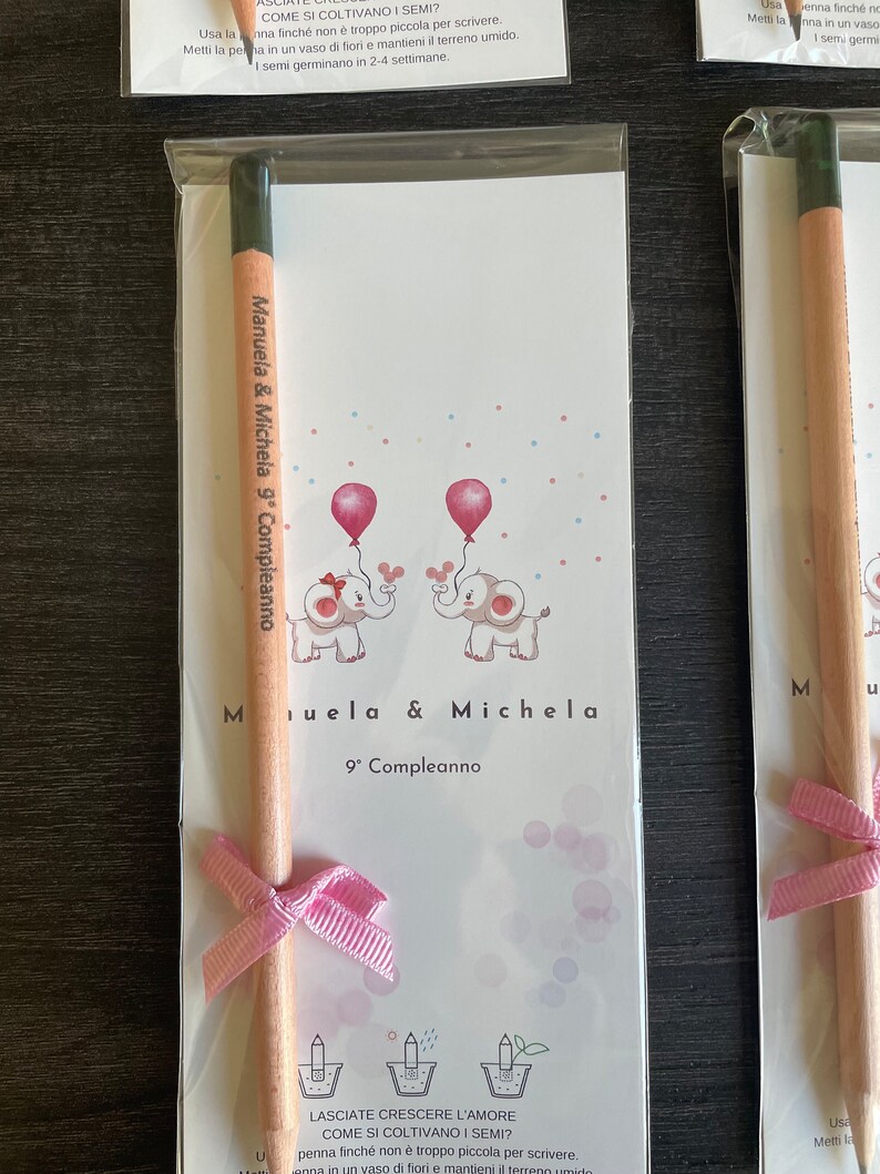 May include: A pink and white card with two elephants holding balloons. The card says "Manuela & Michela 9th Birthday". A wooden pencil with a pink ribbon is attached to the card.