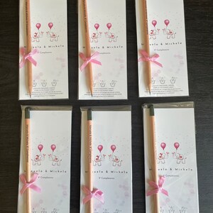 May include: Six pink and white favor boxes with a pencil tied with a pink ribbon. Each box features a cartoon image of two elephants holding balloons and the text "Manuela & Michela 9 Compleanno".