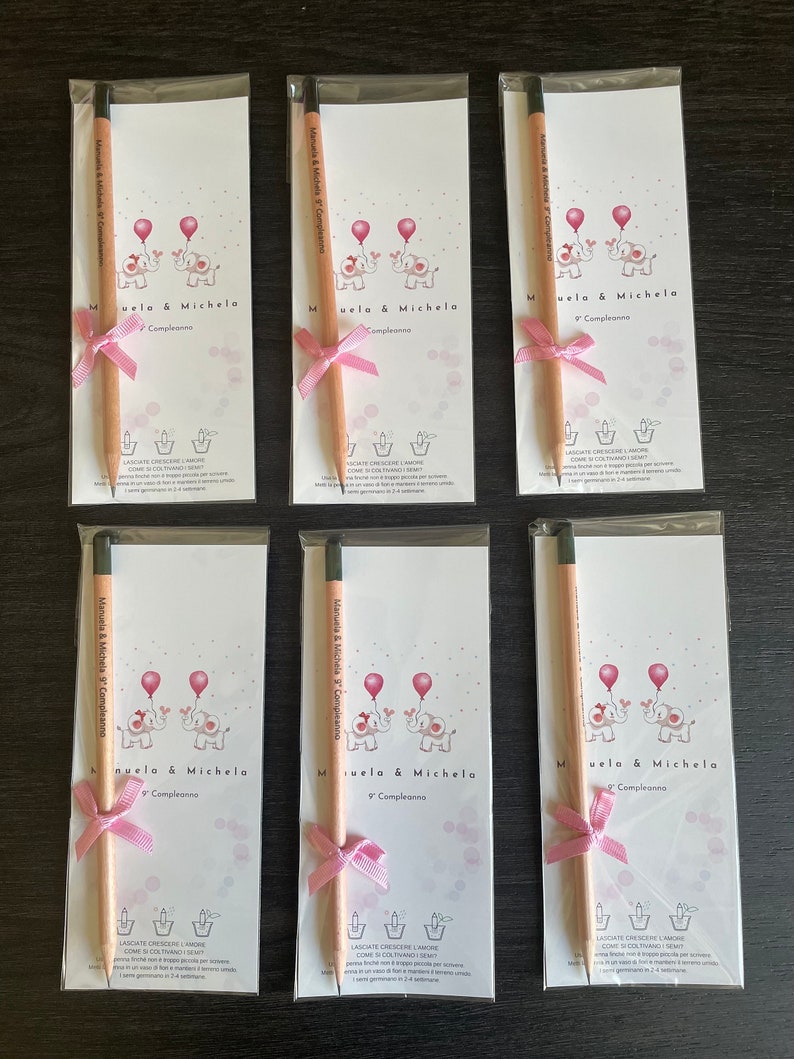 May include: Six pink and white favor boxes with a pencil tied with a pink ribbon. Each box features a cartoon image of two elephants holding balloons and the text "Manuela & Michela 9 Compleanno".