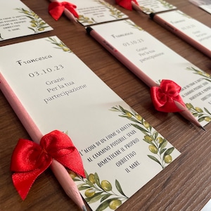 Op de afbeelding: Een roze potlood met een rode strik is bevestigd aan een witte kaart met groene olijftakken en de tekst "Francesca 03.10.23 Grazie Per la tua partecipazione L'acqua di un fiume si adatta al cammino possibile, senza dimenticare il proprio obbiettivo il mare."