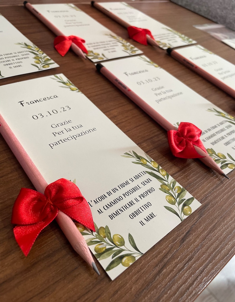Op de afbeelding: Een roze potlood met een rode strik is bevestigd aan een witte kaart met groene olijftakken en de tekst "Francesca 03.10.23 Grazie Per la tua partecipazione L'acqua di un fiume si adatta al cammino possibile, senza dimenticare il proprio obbiettivo il mare."