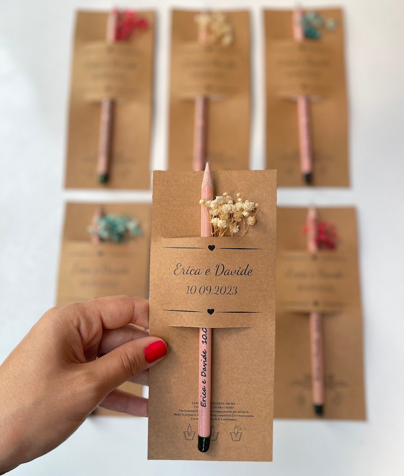 May include: A set of brown cardstock favor boxes with a pink pencil and dried flowers inside. The boxes have the text "Erica e Davide 10.09.2023" printed on them.