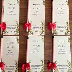 Op de afbeelding: Zes bedankkaartjes met een potlood en een rode strik op elk. De kaartjes zijn groen met olijftakken en de tekst "Francesca 03.10.23 Grazie Per la tua partecipazione" op elk kaartje. De tekst "L'acqua di un fiume si adatta al cammino possibile senza dimenticare il proprio obbiettivo il mare" staat onderaan elk kaartje.