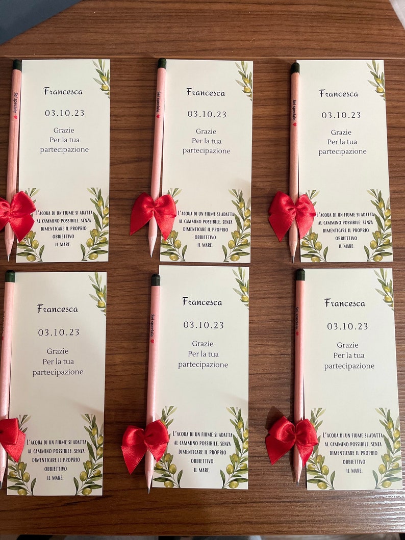 Op de afbeelding: Zes bedankkaartjes met een potlood en een rode strik op elk. De kaartjes zijn groen met olijftakken en de tekst "Francesca 03.10.23 Grazie Per la tua partecipazione" op elk kaartje. De tekst "L'acqua di un fiume si adatta al cammino possibile senza dimenticare il proprio obbiettivo il mare" staat onderaan elk kaartje.