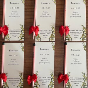Op de afbeelding: Vijf roze potloden met rode strikken die bovenaan zijn gebonden. Elk potlood heeft een wit etiket met de tekst "Francesca" en de datum "03.10.23". De tekst "Grazie Per la tua partecipazione" staat ook op het etiket. De potloden bevinden zich op een witte achtergrond met groene olijftakken en bladeren.
