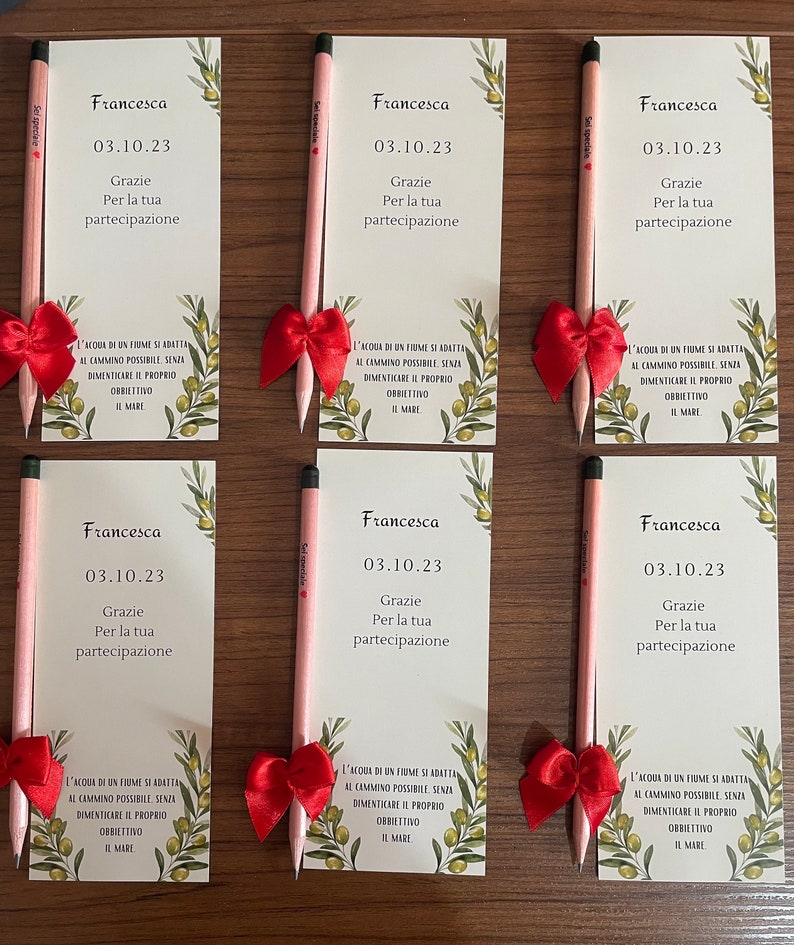 Op de afbeelding: Vijf roze potloden met rode strikken die bovenaan zijn gebonden. Elk potlood heeft een wit etiket met de tekst "Francesca" en de datum "03.10.23". De tekst "Grazie Per la tua partecipazione" staat ook op het etiket. De potloden bevinden zich op een witte achtergrond met groene olijftakken en bladeren.