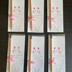 May include: Six white cards with pink and gray illustrations of elephants holding balloons. Each card has a pink ribbon tied around a wooden pencil. The text on the cards reads "Manuela & Michela Compleanno" and "9 Compleanno".