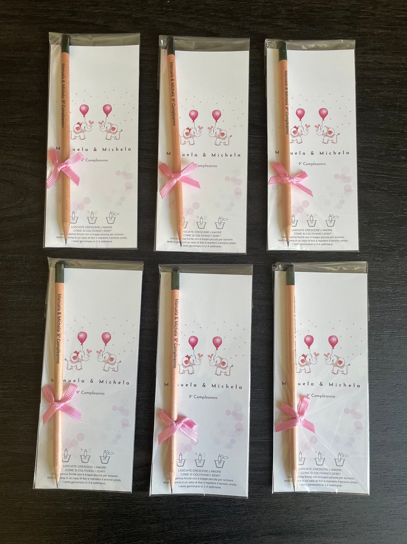 May include: Six white cards with pink and gray illustrations of elephants holding balloons. Each card has a pink ribbon tied around a wooden pencil. The text on the cards reads "Manuela & Michela Compleanno" and "9 Compleanno".