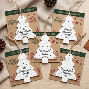 May include: Five white wooden Christmas tree ornaments with jute twine hangers, each featuring black text including family names and the year 2026. The ornaments are displayed on individual kraft paper cards with green headers and Merry Christmas text.