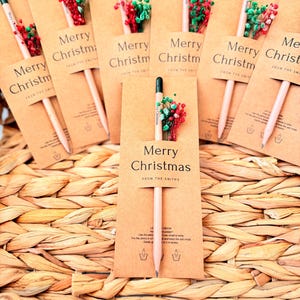 Peut inclure: Plusieurs crayons de Noël avec des sachets de graines, présentés sur un panier en osier. Chaque crayon est accompagné d'une carte en papier kraft imprimée avec "Merry Christmas" et un petit arrangement floral rouge et vert.