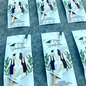 Może przedstawiać: Cztery białe kartki z przymocowanym do każdej oł&oacute;wkiem. Kartki przedstawiają akwarelowe ilustracje dw&oacute;ch os&oacute;b w togach i czapkach absolwent&oacute;w. Tekst na kartkach brzmi "Happy Graduation". Na odwrocie kartek znajdują się instrukcje dotyczące sadzenia oł&oacute;wka, kt&oacute;ry zawiera nasiona. Tekst brzmi "Let Love Grow".