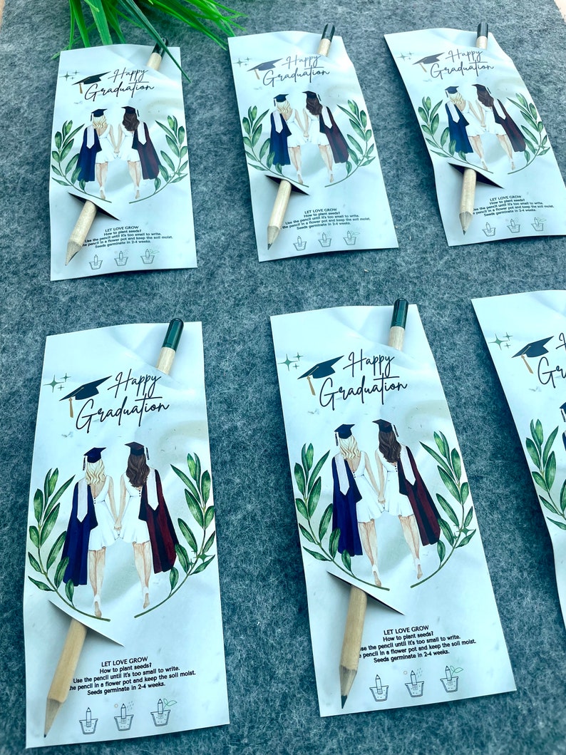 Może przedstawiać: Cztery białe kartki z przymocowanym do każdej oł&oacute;wkiem. Kartki przedstawiają akwarelowe ilustracje dw&oacute;ch os&oacute;b w togach i czapkach absolwent&oacute;w. Tekst na kartkach brzmi "Happy Graduation". Na odwrocie kartek znajdują się instrukcje dotyczące sadzenia oł&oacute;wka, kt&oacute;ry zawiera nasiona. Tekst brzmi "Let Love Grow".