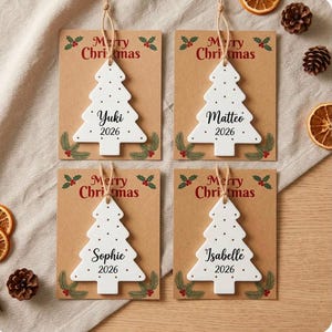 May include: Four white ceramic Christmas tree ornaments with jute twine hangers, each mounted on a brown card printed with Merry Christmas, holly, and the names Yuki, Matteo, Sophie, or Isabelle above the year 2026 in black script and font.