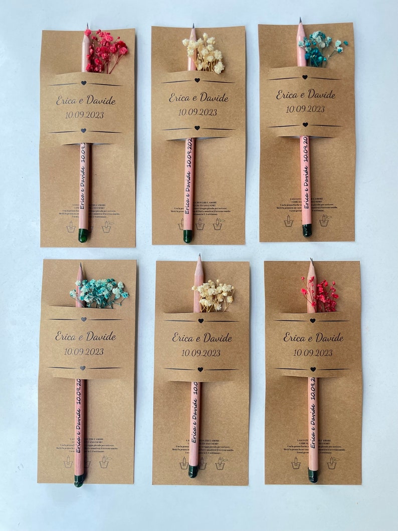 May include: Six kraft paper favor boxes with pink pencils and dried flowers. Each box has the text "Erica & Davide" and the date "10.09.2023".