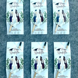 Może przedstawiać: Sześć białych kopert z papieru z oł&oacute;wkiem w środku każdej. Koperty mają zielony wz&oacute;r liści i rysunek dw&oacute;ch kobiet w togach absolwent&oacute;w. Tekst na kopertach brzmi "Happy Graduation".